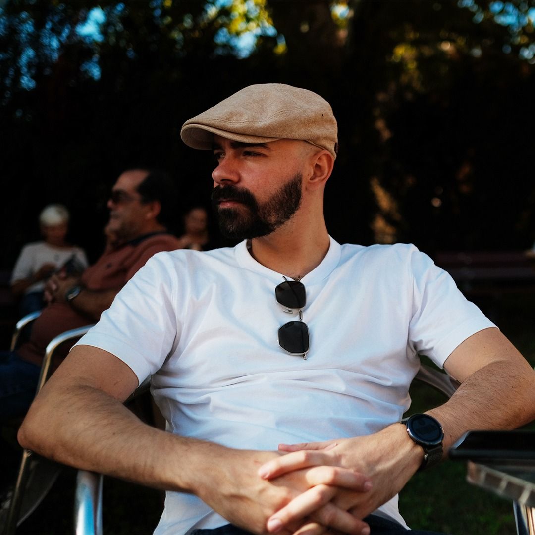Autoportrait d’Eyekust en casquette beige et t-shirt blanc, assis en terrasse, le regard tourné sur le côté.